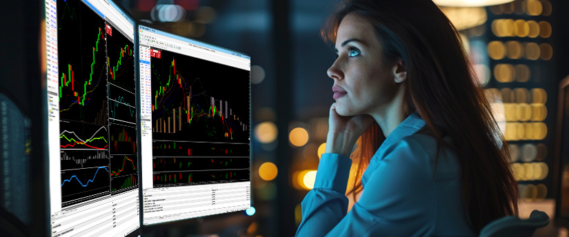 A female trader looking closely at financial candlestick charts and bid-ask spreads in trading across multiple monitors at night.